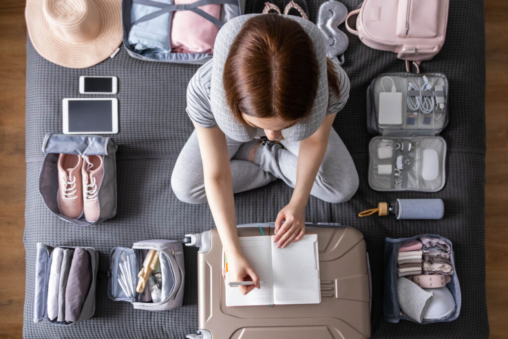 Smiling tourist woman packing suitcase to honeymoon in April writing paper list sitting on bed at home. Female getting ready to travel trip organization things storage in comfy cases bags use konmari method