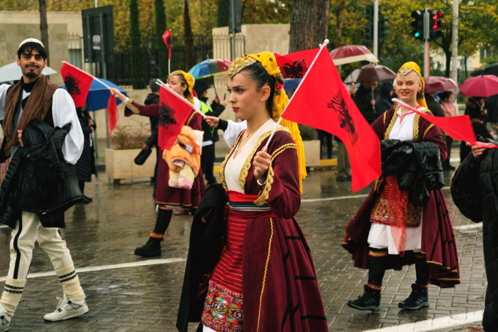 Tirana, Albania - November 28, 2023: On Avenue of Martyrs of the Nation, students and people in traditional attire enthusiastically march with Albanian flags on a rainy Independence Day, with spectators holding umbrellas and flags in the background.