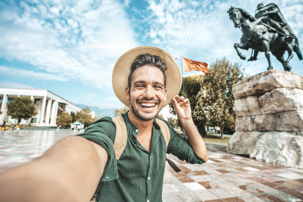 Happy tourist taking selfie at Skanderbeg square in Tirana, Albania - Smiling handsome young man laughing at camera outside - Summer vacation and European holidays concept